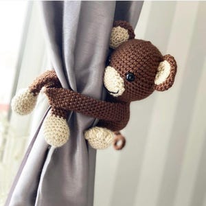 May include: A brown and beige crocheted monkey curtain tieback. The monkey is hugging a gray curtain, with its arms wrapped around the fabric. The monkey has black eyes and a smiling face. The curtain tieback is a decorative item.