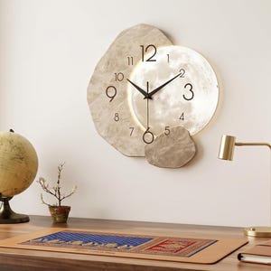 May include: A wall clock featuring a circular face with black numerals and hands, partially overlaid by an irregular, textured beige stone-like frame. The clock design incorporates a smooth, metallic-toned circular section behind the hands.