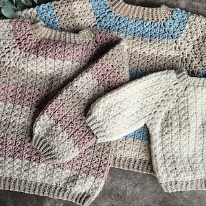May include: Three handmade crochet sweaters in various sizes. The sweaters feature a textured stitch pattern and are in neutral colors with stripes of pink, blue, and cream. The sleeves and necklines have a ribbed design.