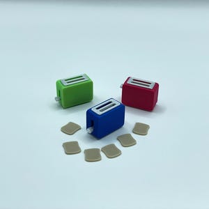 Op de afbeelding: Drie miniatuur speelgoedtoasters in groen, rood en blauw, elk met een zilveren bovenkant en een kleine hendel. Verschillende kleine, bruinachtige toastschijfjes zijn rond de toasters op een wit oppervlak gerangschikt.