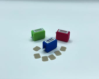 Può includere: Tre tostapane giocattolo in miniatura verdi, rossi e blu, ciascuno con una parte superiore argentata e una piccola leva. Diverse piccole fette di pane tostato color beige sono disposte attorno ai tostapane su una superficie bianca.