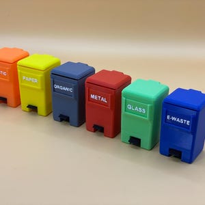 Puede incluir: Siete contenedores de reciclaje en miniatura de diferentes colores, etiquetados con los siguientes tipos de residuos: Plástico, Papel, Orgánico, Metal, Vidrio, Residuos electrónicos.