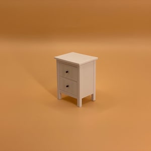 May include: A small, white bedside table with two drawers and black knobs. The table has a simple, rectangular design and stands on four short legs. The piece is set against a solid, light brown background.
