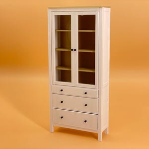 May include: White wooden cabinet with glass doors and three drawers. The cabinet has a light wood top and black knobs.