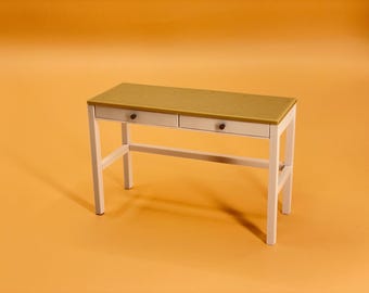 Könnte beinhalten: Ein weißer Schreibtisch mit zwei Schubladen und einer hellbraunen Tischplatte. Der Schreibtisch hat ein schlichtes Design und ist aus Holz.