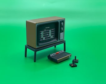 May include: A miniature retro television set with a brown cabinet and a black screen displaying a video game. The television is on a small black table. There is a vintage video game console and two controllers on the green surface.