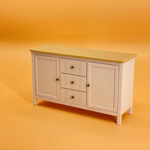 Puede incluir: Un aparador blanco con tapa amarilla y pomos negros. El aparador tiene dos puertas y tres cajones. El aparador está sobre un fondo amarillo.