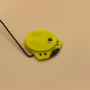 May include: A bright yellow tape measure with a black measuring tape and a black wire. The tape measure has a rounded shape with a small hole and three smaller holes. The background is a solid tan color.