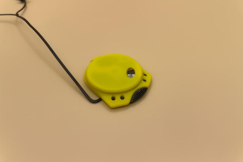 May include: A bright yellow tape measure with a black measuring tape and a black wire. The tape measure has a rounded shape with a small hole and three smaller holes. The background is a solid tan color.