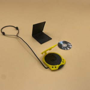 May include: A miniature yellow and black portable CD player with a CD, a black CD case, and black headphones. The CD player has a circular design with a yellow lid and a black center. The headphones are connected to the CD player.