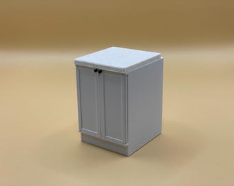 May include: A small, light gray cabinet with two doors and a white speckled countertop. The cabinet has black knobs and a simple, modern design. The background is a solid, light tan color.