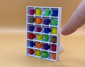 Può includere: Una piccola mensola bianca piena di piccole tazze colorate. Le tazze sono di vari colori, tra cui rosso, giallo, verde, blu, arancione e viola. La mensola è su uno sfondo arancione chiaro.