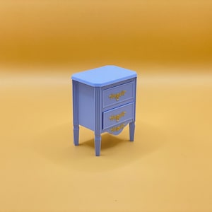 Puede incluir: Una pequeña mesita de noche de madera azul claro con dos cajones y herrajes de color dorado. La mesita de noche tiene una base ligeramente curvada y se encuentra sobre una superficie amarilla.