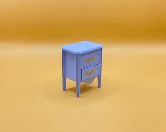 Può includere: Un piccolo comodino in legno blu chiaro con due cassetti e maniglie dorate. Il comodino ha una base leggermente curva e si trova su una superficie gialla.