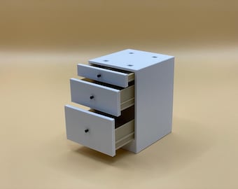 May include: A small, white, three-drawer cabinet with black knobs. The drawers are partially open, revealing the interior. The cabinet is set against a light tan background. The top of the cabinet has four small square indentations.