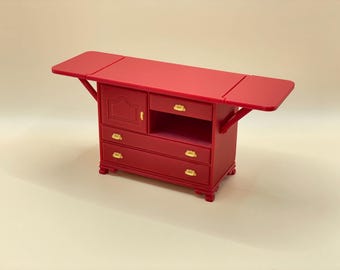 Può includere: Un mobile in legno rosso con tre cassetti e una porta. Il mobile ha un piano da tavolo ribaltabile.