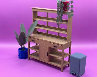 Può includere: Una credenza in legno marrone con uno scaffale in cima e un armadio con cassetti e ripiani sotto. La credenza è decorata con una pianta in vaso con foglie verdi e un piccolo bidone della spazzatura.