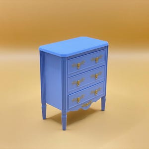 Puede incluir: Una pequeña cómoda azul claro con tres cajones y herrajes de color dorado. La cómoda tiene una parte inferior ligeramente curvada y se apoya en cuatro patas.
