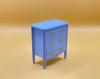 Può includere: Un piccolo comò blu chiaro con tre cassetti e maniglie dorate. Il comò ha un fondo leggermente curvo e poggia su quattro gambe.