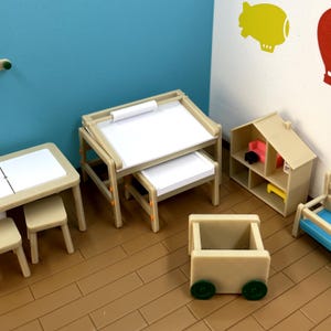 Puede incluir: Un conjunto de muebles de madera en miniatura para una casa de muñecas, que incluye un escritorio con una tapa blanca, una silla, una estantería, una casa de muñecas y un carro de juguete. Los muebles están pintados de color marrón claro con detalles en naranja.