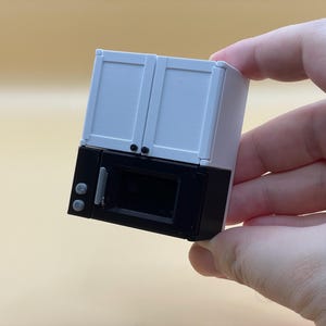 Può includere: Un mobile da cucina in miniatura e combinazione microonde. Le ante superiori dell'armadio sono bianche e il microonde sottostante è nero. Il piccolo elettrodomestico è tenuto in mano su uno sfondo neutro.