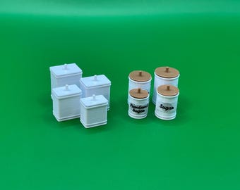 Può includere: Set di sei barattoli in ceramica bianca con coperchi. Quattro barattoli sono quadrati e due sono rotondi. I barattoli rotondi hanno coperchi marroni e sono etichettati "Zucchero" e "Zucchero a velo".