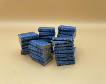 Può includere: Pile di libri blu in miniatura con bordi dorati e la parola "Britannica" stampata sul dorso. I libri sono disposti in diversi mucchi su uno sfondo neutro.