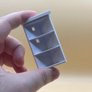 May include: White miniature bookshelf with three shelves and a small rectangular opening on each shelf.