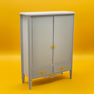 May include: A gray wooden cabinet with two doors and two drawers. The cabinet has gold-colored hardware and decorative details on the bottom.