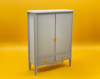 Può includere: Un armadio in legno grigio con due ante e due cassetti. L'armadio ha maniglie dorate e dettagli decorativi nella parte inferiore.