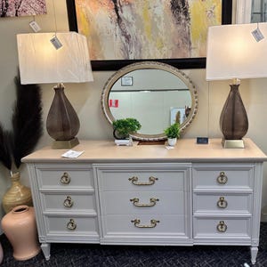 May include: A white dresser with six drawers and gold hardware. The dresser has a light wood top and is decorated with two lamps with white shades and gold bases. A round mirror with a gold frame is centered above the dresser.