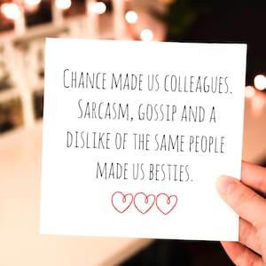 May include: A white card with black text that reads "Chance made us colleagues. Sarcasm, gossip and a dislike of the same people made us besties." There are three red hearts at the bottom of the card.