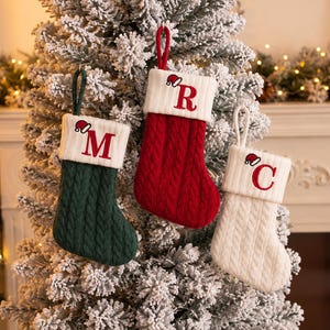 May include: Three cable-knit Christmas stockings with white cuffs. The left stocking is dark green with the letter M, the center is red with the letter R, and the right is white with the letter C. Each cuff features a small embroidered Santa hat.