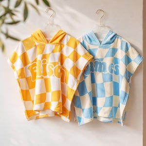 May include: Two hooded beach ponchos hanging on clear hangers. One is yellow and white checkered with the word "Elisa" in orange. The other is blue and white checkered with the word "James" in blue. Both are made of a soft, absorbent material.