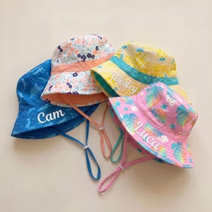 May include: Four patterned bucket hats, each with a name. A blue hat has a dinosaur design and the name "Cam". A floral hat has a pink band. A yellow pineapple-print hat says "Wesley". A pink hat has a leaf pattern and the name "Luca".