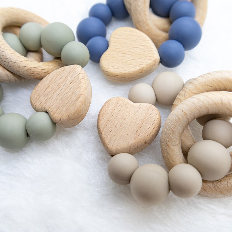 May include: Three wooden teething rings with silicone beads in shades of blue, green, and beige. The rings are shaped like circles and hearts.