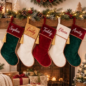 May include: Five velvet Christmas stockings hanging in a row, featuring deep green, gold, and red colours. Each stocking has a contrasting cuff embroidered with the names Sophia, Emily, Teddy, Olivia, and Amity, finished with gold tassels.