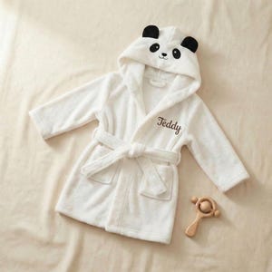 May include: White hooded baby bathrobe with a panda face design on the hood. The robe has a matching belt, pockets, and the name "Teddy" embroidered on the front. A wooden rattle is next to the robe.