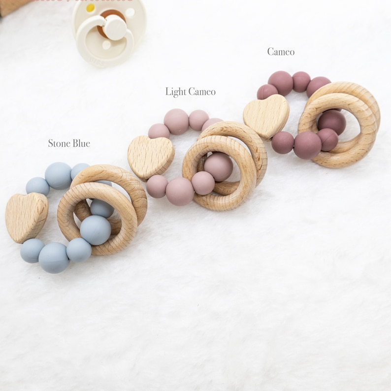 May include: Three wooden teething rings with silicone beads in different colours: stone blue, light cameo, and cameo. Each ring has a wooden heart attached.