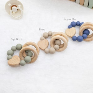 May include: Four wooden teething rings with silicone beads in different colours: sage green, taupe, Aegean blue. Each ring has a wooden heart charm.