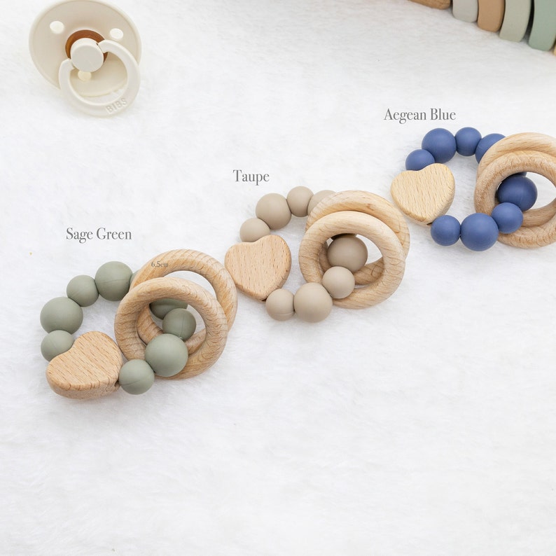 May include: Four wooden teething rings with silicone beads in different colours: sage green, taupe, Aegean blue. Each ring has a wooden heart charm.