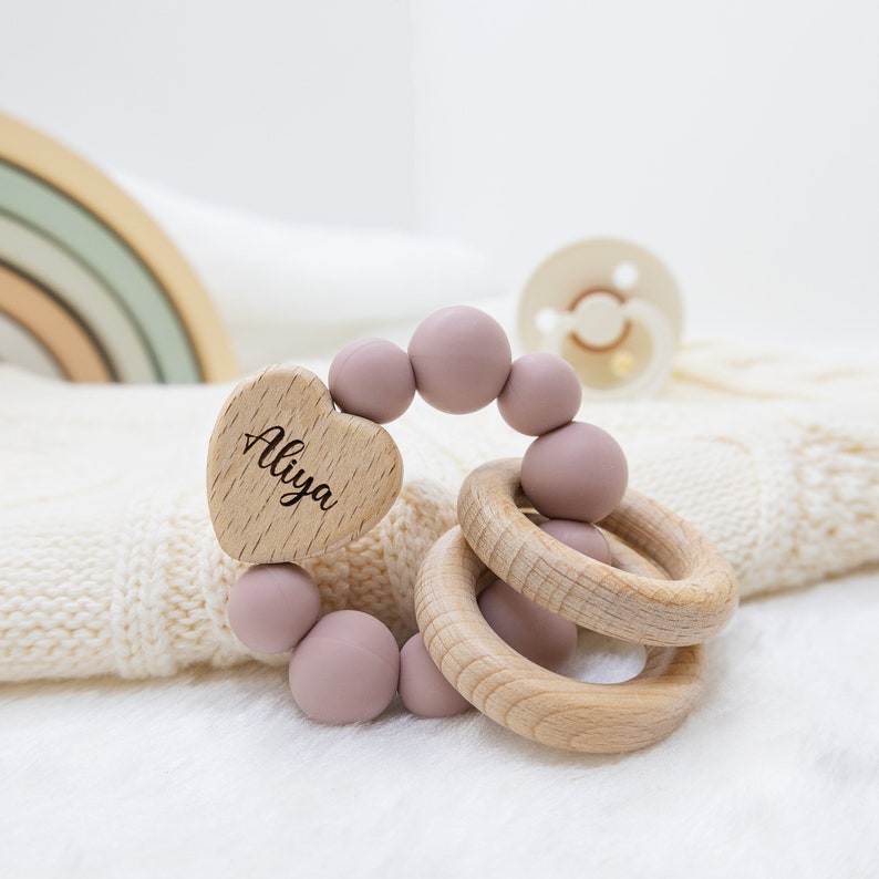 May include: A pink and wooden baby teether with a heart-shaped wooden tag that says "Aliya".