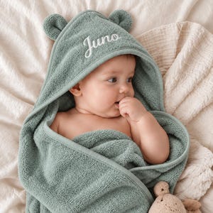 Peut inclure: Une serviette de bain à capuche pour bébé de couleur vert sauge, dotée de petites oreilles d'ours arrondies sur la capuche. Le prénom Juno est brodé en lettres cursives blanches sur le devant. La serviette est en tissu éponge texturé.