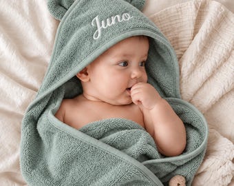 Peut inclure: Une serviette de bain à capuche pour bébé de couleur vert sauge, dotée de petites oreilles d'ours arrondies sur la capuche. Le prénom Juno est brodé en lettres cursives blanches sur le devant. La serviette est en tissu éponge texturé.