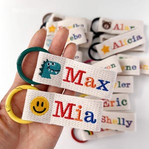 May include: A collection of personalised fabric keychains made of off-white woven material. Each keychain features a colourful embroidered name and a small icon, such as a dinosaur or smiley face, attached to a round plastic ring in various colours.