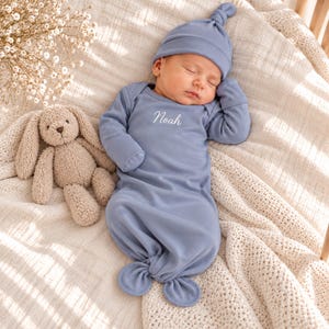 May include: A sleeping infant wearing a dusty blue long-sleeve knotted gown and a matching knotted hat. The gown features the name Noah embroidered in white script on the chest. A beige plush bunny toy is positioned next to the infant.