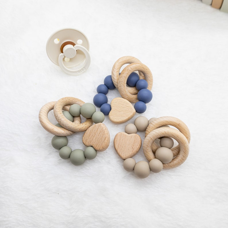 May include: Three wooden teething rings with silicone beads in blue, green, and beige. Each ring has a wooden heart attached.
