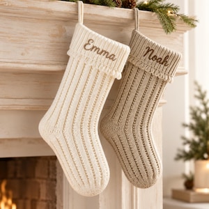 May include: Two knitted Christmas stockings hanging side by side. The left stocking is cream-coloured with the name Emma embroidered in brown. The right stocking is tan with the name Noah embroidered in brown. Both feature a ribbed texture and fringe.