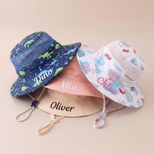 Puede incluir: Cuatro sombreros de sol personalizados. Uno es azul marino con un estampado de dinosaurios y el nombre "Dino". Otro es rosa con "Mia". Un tercero es beige con "Oliver". El último es blanco con un estampado de flamencos y "Chloe".