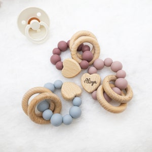 May include: Three wooden teething rings with silicone beads in blue and pink. The rings are circular and have a heart-shaped wooden bead with the name "Aliya" engraved on it.